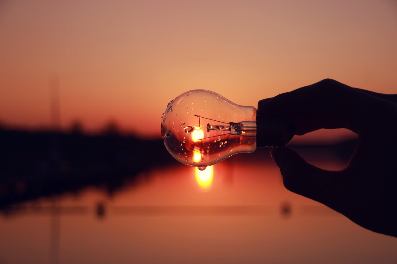 hero-img-02 Silhouette of a hand holding a light bulb against a stunning sunset over a lake, blending creativity and nature.